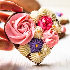 someone is holding a heart shaped cake with flowers on it's side and another hand over the top