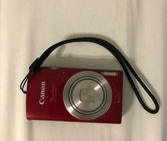 a red digital camera sitting on top of a white bed next to a black cord