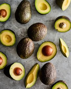 an avocado cut in half and whole on a gray surface with other fruits