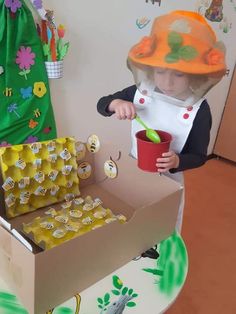 a child in an orange hard hat is holding a cup and spoon while standing next to a cardboard box filled with eggs