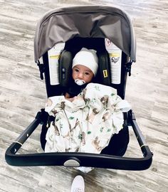 a baby sitting in a stroller wearing a white hat and blanket with an infant pacifier in it's mouth