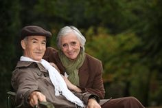 an elderly couple sitting on a bench in the park, looking at the camera and smiling