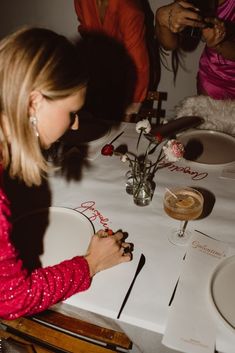 a woman sitting at a table writing on a piece of paper with flowers in the background