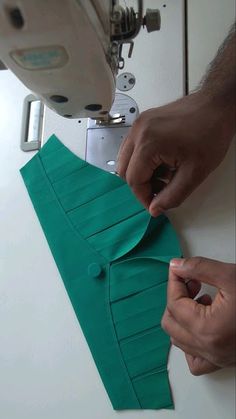 two hands are working on a green piece of paper with a sewing machine in the background