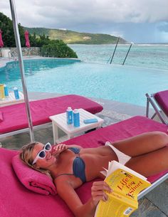 a woman laying on top of a pink towel next to a pool reading a book