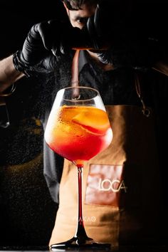 a person in an apron is pouring a drink into a wine glass with orange and red liquid