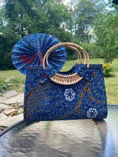 "Stand out with this beautiful, vibrant blue Dakashi African wax print bag! The bold design is sure to make any outfit pop!  This is a large-sized bag with a large carrying capacity and with a hardboard shaping the entire purse, it is extra sturdy. There is a black lining, and interior zipper to securely close the bag. The straps are woven from wood cane. It also comes with a large, matching, foldable hand fan made of cloth and leather.  The approximate dimensions of the bag are: 15\" wide 11.5\ Blue Handheld Shoulder Bag, Traditional Blue Shoulder Bag For Daily Use, Traditional Blue Shoulder Bag For Everyday Use, Traditional Blue Shoulder Bag For Everyday, Traditional Blue Bag For Daily Use, Traditional Blue Bags For Daily Use, Traditional Blue Rectangular Shoulder Bag, Traditional Blue Rectangular Bag, Blue Bags With Braided Handles