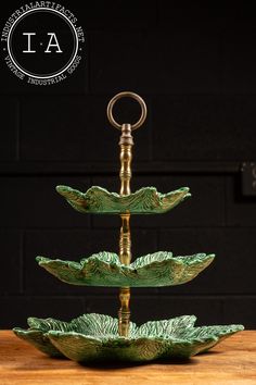 three tiered green glass bowl with metal handles on a wooden table top in front of a black brick wall