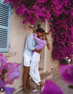 a man and woman are kissing in front of purple bougaia flowers on the side of a building