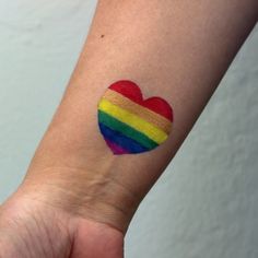 a small rainbow heart tattoo on the left inner arm, with a red and yellow stripe across it