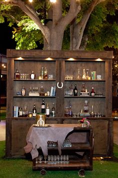 an outdoor bar set up with bottles and glasses on the table in front of a tree