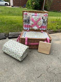 a wicker picnic basket with pink ribbon and matching cushions sits on the ground next to two pieces of luggage