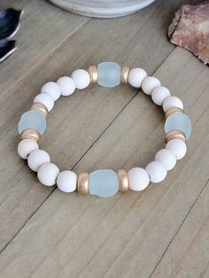 This stacker boasts beautiful white wooden and gold beads, alongside environmentally friendly recycled glass from the Krobo tribe. Embrace a boho and neutral aesthetic while making a positive impact with this stylish piece.    Cream colored wooden beads   Gold heishi colored beads   Aqua Recycled glass   Fits a wrist up to 6.5"   Created in studio - Wilmington, NC White Wooden Beads Bracelet, White Recycled Glass Jewelry For The Beach, White Stretch Bracelet With Round Wooden Beads, White Bracelets With Round Wooden Beads, Wood Bead Bracelet, Neutral Aesthetic, Wood Bracelet, Gold Armband, Wilmington Nc