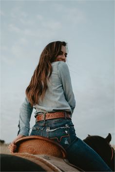 a woman sitting on the back of a horse wearing jeans and a cowboy hat with her hair blowing in the wind