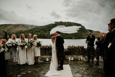 a bride and groom kissing in front of their wedding party on the shores of a lake