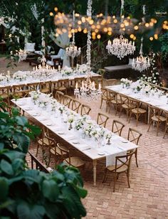 an outdoor dining area with long tables and chandeliers hanging from the ceiling, surrounded by greenery