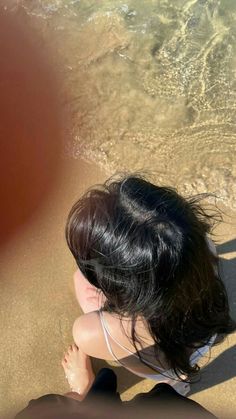 a woman sitting on top of a sandy beach next to the ocean with her hair blowing in the wind