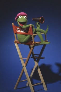 kermie the frog sitting in a wooden folding chair with a red hat on