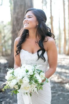 a woman in a wedding dress holding a bouquet and smiling at the camera with trees behind her