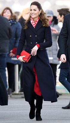 a woman in a black coat and red dress is walking down the street with other people behind her