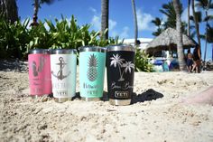 four different colored cups are sitting in the sand near palm trees and people on the beach