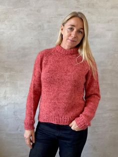 a woman standing in front of a wall wearing a red knitted sweater and jeans