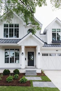 a white house with two story windows and a black door on the front porch is surrounded by greenery