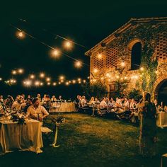 a group of people sitting at tables in front of an old building with lights strung from the ceiling