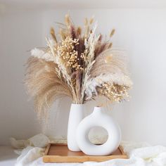 a white vase with dried plants in it sitting on a wooden tray next to a blanket