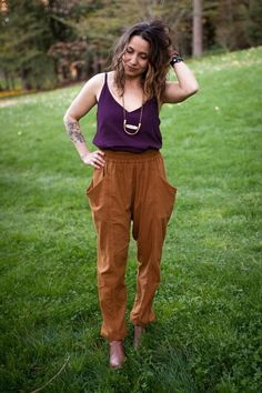 a woman standing in the grass with her hands on her head and wearing brown pants