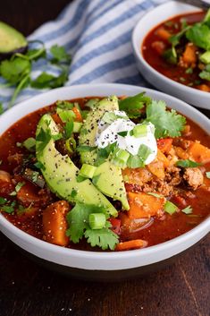 two white bowls filled with chili and meat soup on top of a wooden table next to an avocado