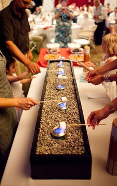 people standing around a long table with candles on it and one person lighting the candles