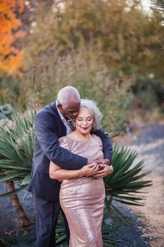 an older man and woman embracing each other in front of some trees with orange leaves