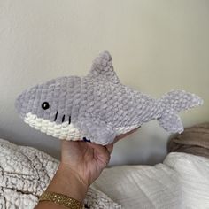 a hand holding up a knitted shark toy in front of a white wall background