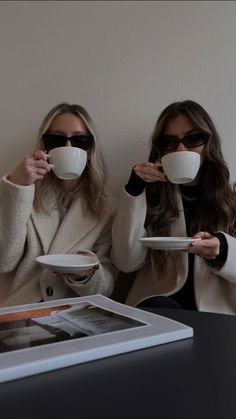 two women sitting at a table with coffee cups in front of their faces, both holding plates