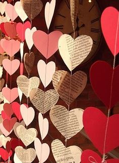 many heart shaped paper hanging from strings on a wall with a clock in the background