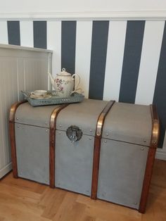 an old trunk is sitting on the floor in front of a striped wall with a teapot and tray