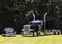 two old cars are parked next to a large truck on the grass in front of some trees