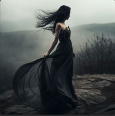 a woman standing on top of a rocky hillside wearing a black dress and flowing hair