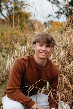 a young man sitting on the ground in front of some tall grass and smiling at the camera