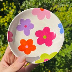 a hand holding a bowl with flowers painted on it in front of some trees and bushes