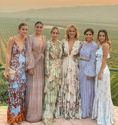 four women in dresses posing for a photo at an outdoor event with vineyards in the background