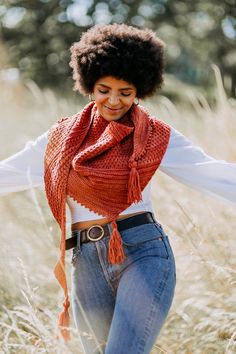 a woman is walking through tall grass wearing a red scarf and jeans with her hands in her pockets