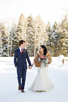 a bride and groom holding hands in the snow with pine trees in the back ground