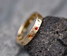 a gold wedding band with red and blue stones on top of a rock in front of some rocks