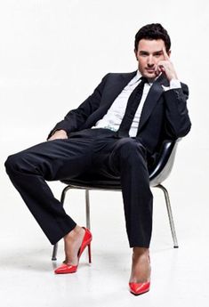 a man in a suit and red shoes sitting on a chair with his hand to his face