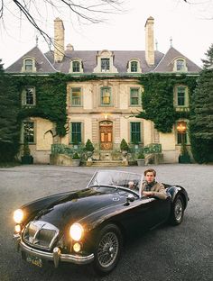 a man sitting in an old fashioned sports car with ivy growing on the building behind him