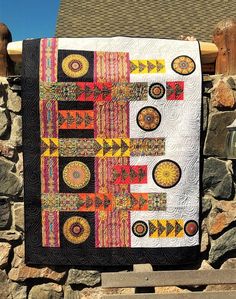 a quilt hanging on a stone wall next to a wooden fence with a building in the background