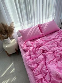 a bed with pink sheets and pillows in front of a white curtained window next to a plant