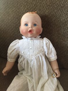 an old fashioned baby doll with blue eyes and white dress sitting on the floor in front of a couch